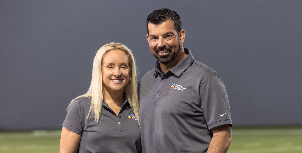 Nina and Ryan Day stand together on a football field wearing Kids Mental Health Foundation polos, representing their support for youth mental health initiatives.
