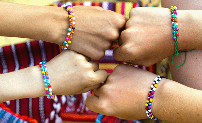 Close-up of five hands wearing colorful beaded friendship bracelets, joined together in a fist bump over a vibrant woven background, symbolizing unity, creativity, and youth connection.