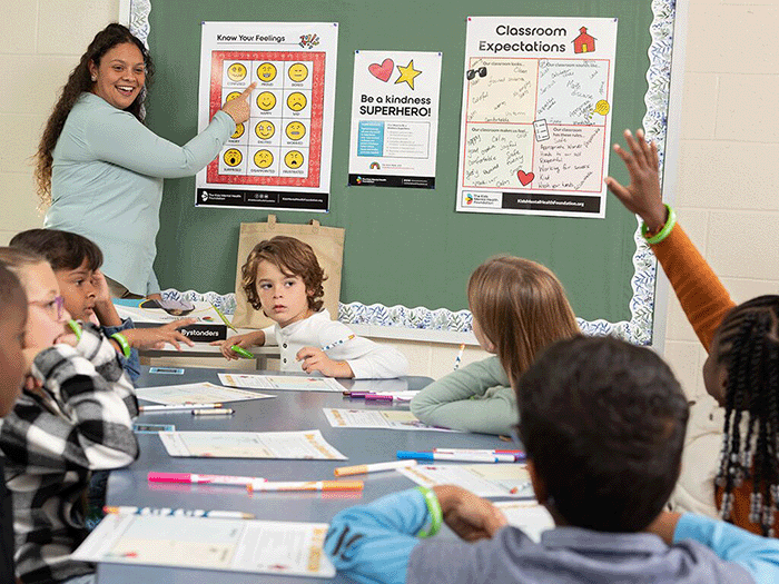 Elementary school teacher leads a classroom activity on emotional awareness using a 'Know Your Feelings' chart, as students engage with worksheets and raise their hands, promoting social-emotional learning through The Kids Mental Health Foundation's Teacher Kit.