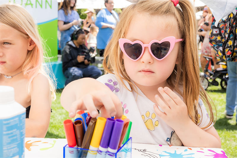 Young girl in heart-shaped sunglasses selects a marker at a kids’ craft table during a community event, representing hands-on activities to support youth engagement and mental wellness.